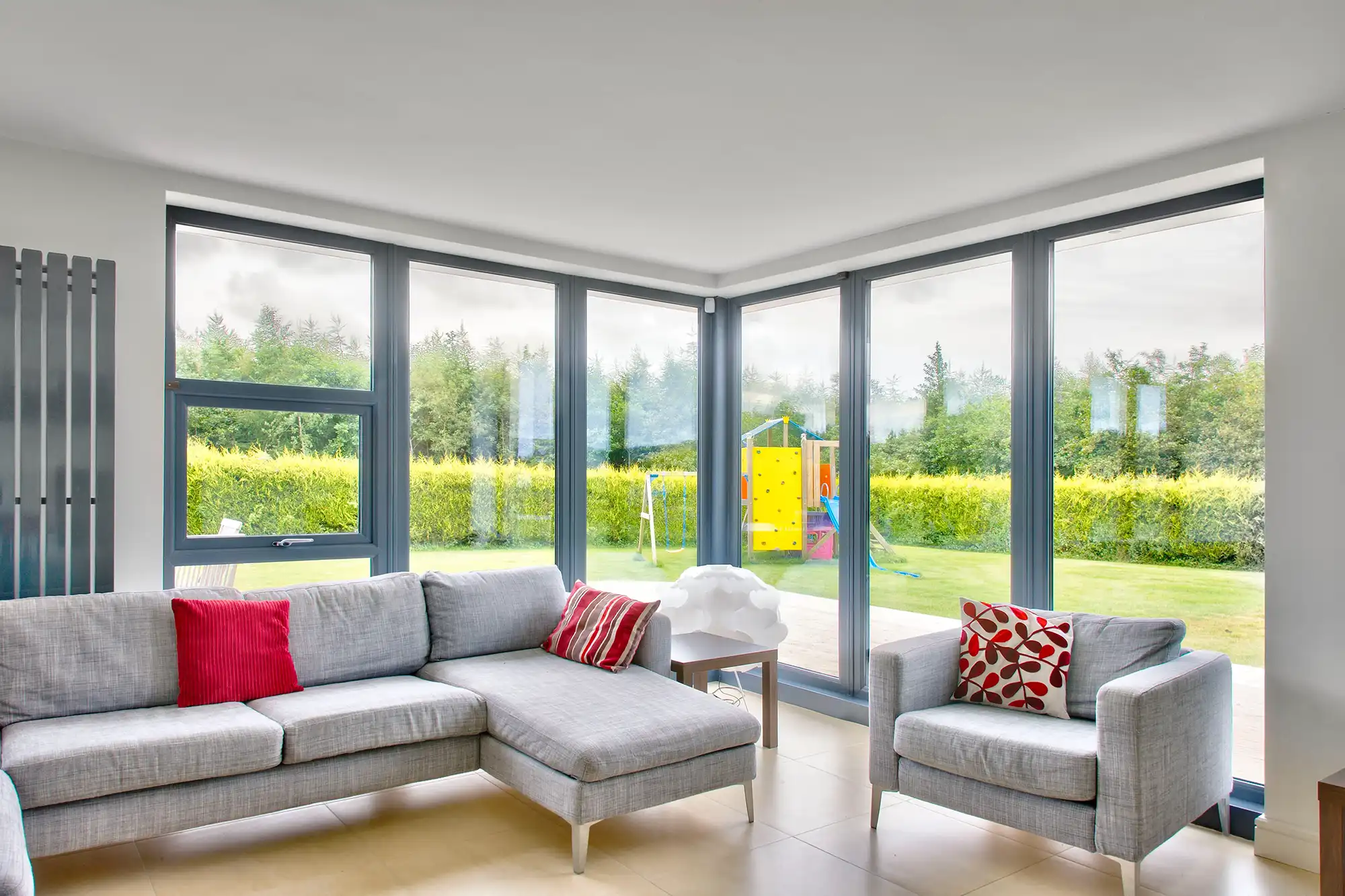 Modern living room with large windows, gray furniture, and a view of a garden featuring play equipment. Bright and airy atmosphere.