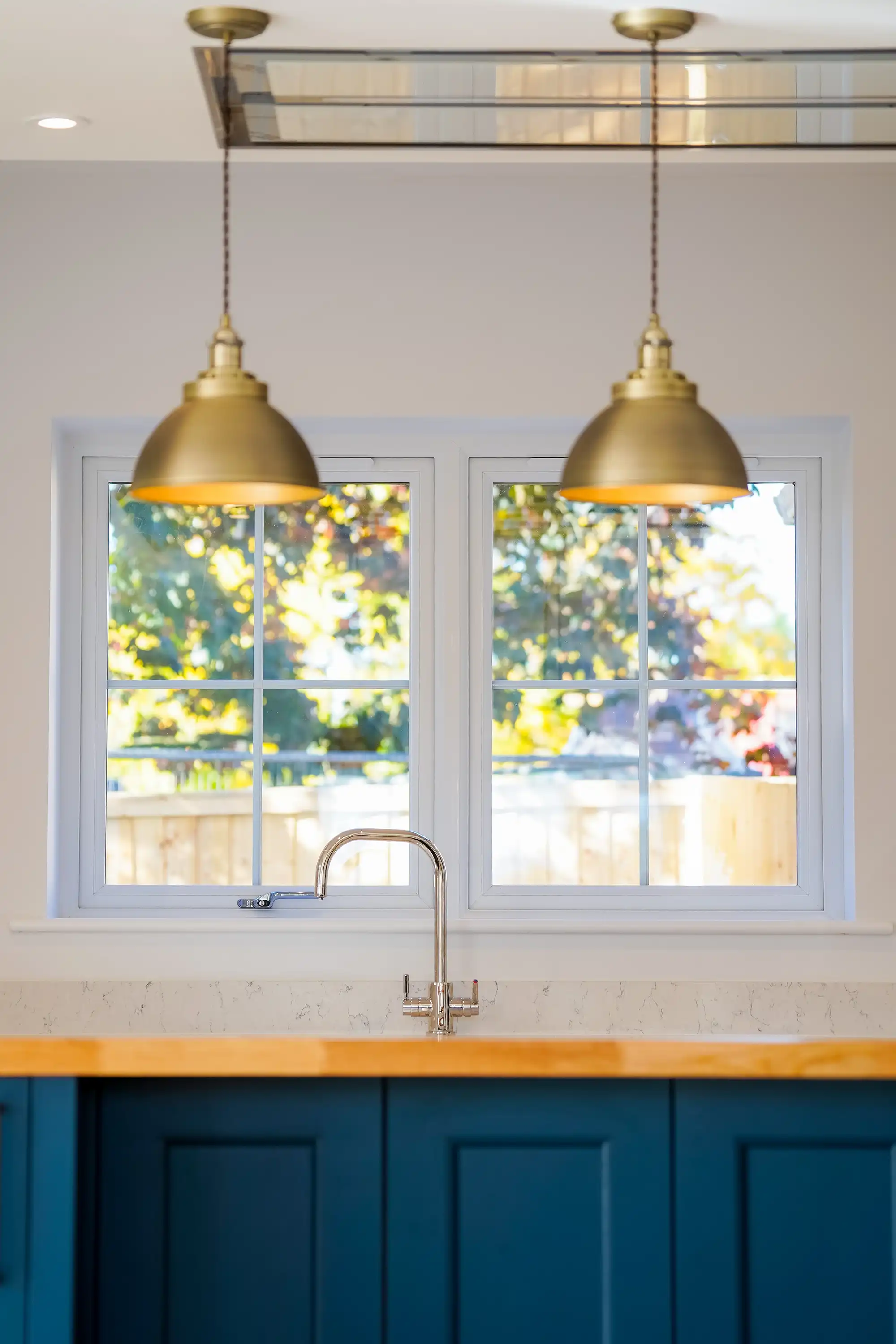 Bright kitchen with a wooden countertop, blue cabinetry, and elegant brass pendant lights above a modern sink and windows overlooking greenery.