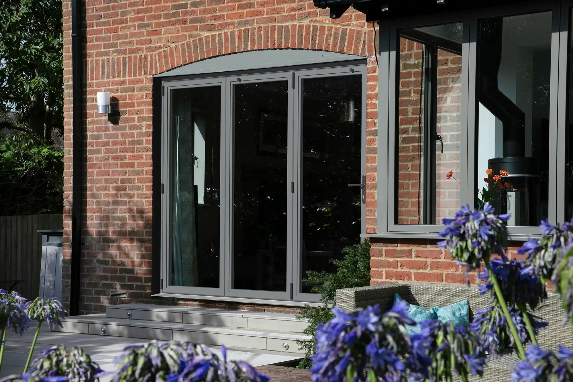 A grey multi-panel sliding door on a brick house, with flowers in the foreground and a serene outdoor seating area.