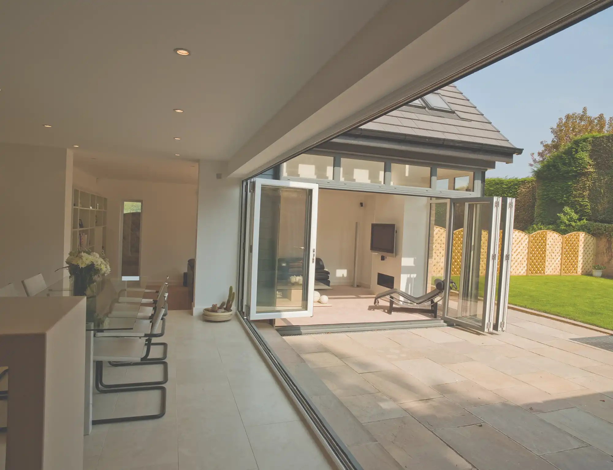 Modern open-plan living area with glass doors leading to a garden. Features a dining table and lounge space, bright and airy design.