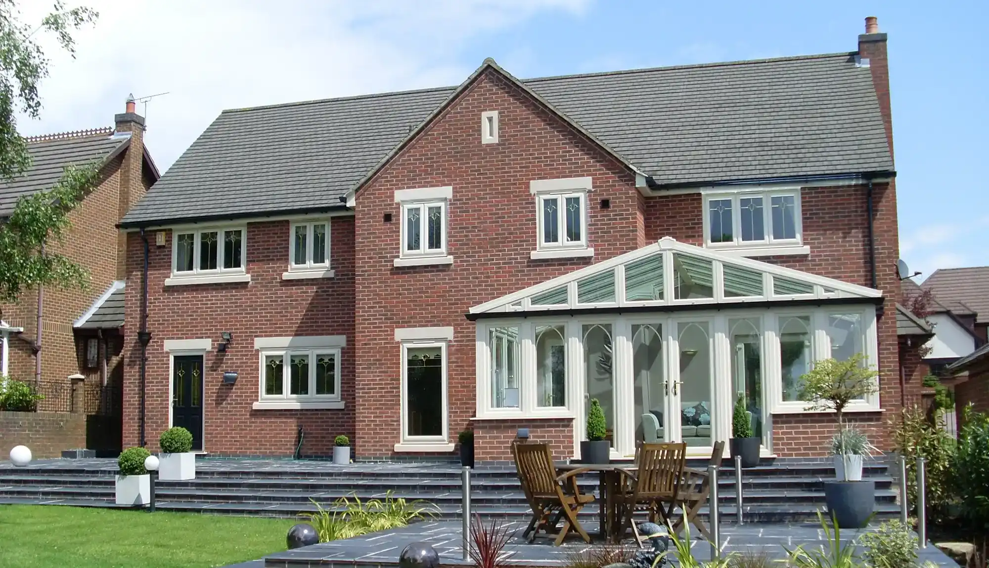 A modern brick house with a conservatory, lush lawn, and patio furniture, set against a clear blue sky.